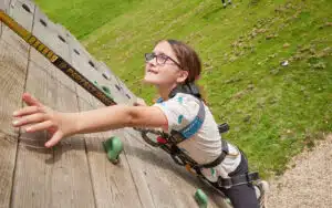 A child wearing glasses and a harness climbs a wooden outdoor climbing wall, reaching up with one hand. Grass and gravel are visible below.