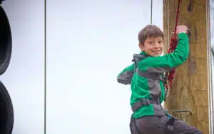 A boy in a green jacket and harness climbs a wooden pole during an outdoor ropes course activity, smiling at the camera.