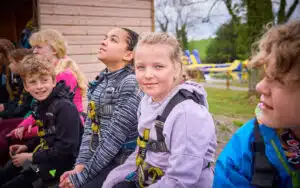 A group of children wearing harnesses sit outside near a wooden building, with inflatable water equipment visible in the background.