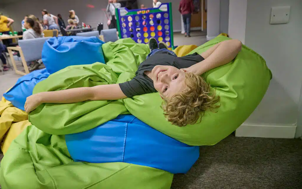 A child lies on a pile of green and blue bean bags, smiling at the camera. People in the background play games in a casual indoor setting.