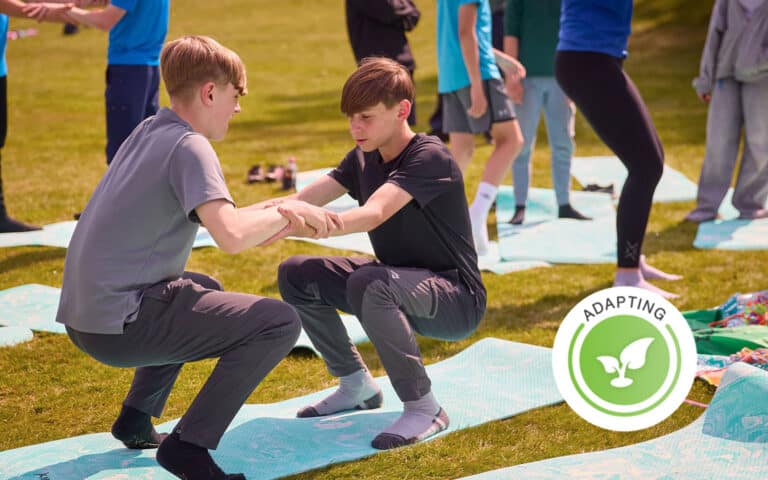 Two boys balance facing each other on exercise mats, holding hands, while others engage in multi-activity exercises in the background on grass. A circular "Adapting" symbol is in the lower right corner.