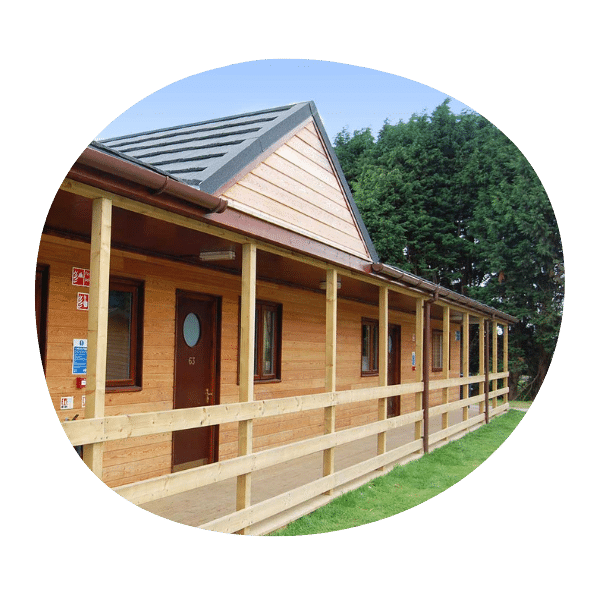 A single-story wooden building with a covered porch and railing, set on a grassy area with trees in the background.