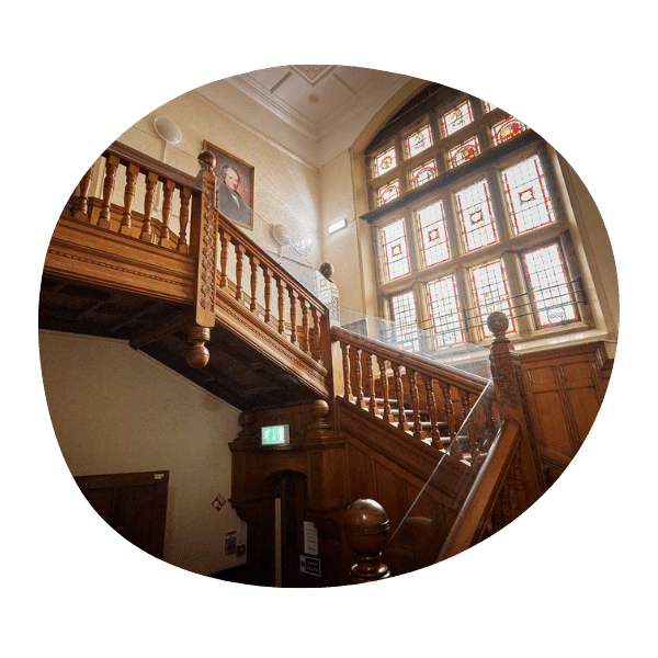 Wooden staircase with carved railings in a historic building, featuring large stained glass windows and a portrait hanging on the wall.