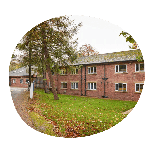 A two-story brick building with multiple windows is surrounded by grass, trees, and autumn leaves on the ground.