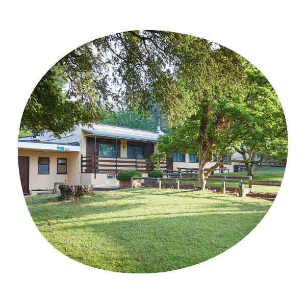 Single-story building with a front porch and railing, surrounded by trees and a grassy lawn, photographed during daylight.