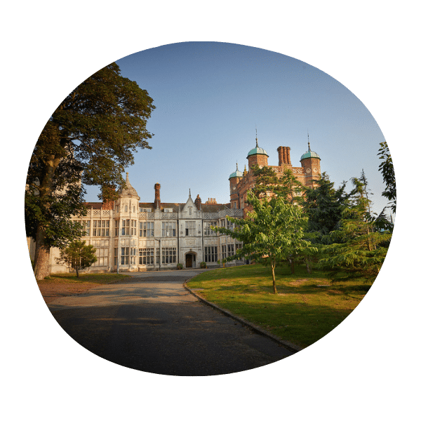 A grand historic building with multiple turrets, large windows, and a tree-lined driveway under a clear sky.