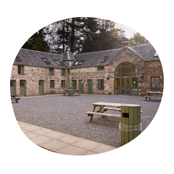 A stone building with green doors and windows surrounds a gravel courtyard with picnic tables and a green trash bin, set against a backdrop of tall trees.