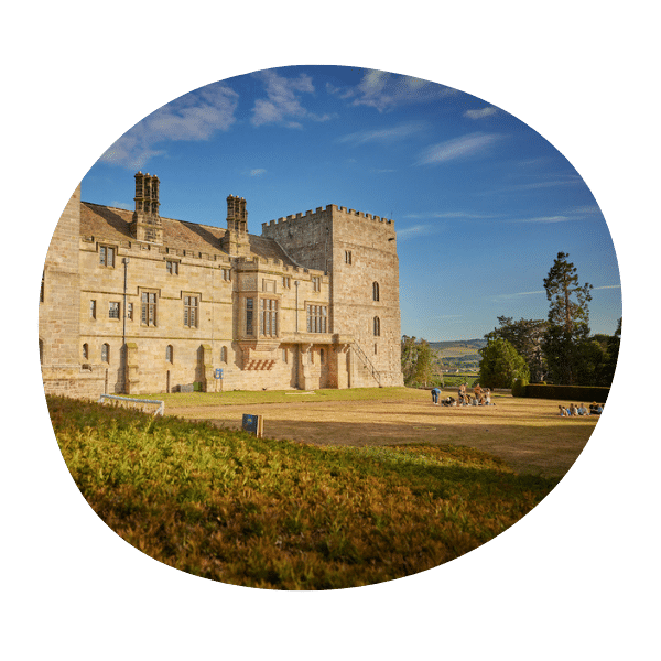 A historic stone castle with a square tower stands under a blue sky, surrounded by grass and a few trees, with distant hills visible in the background.