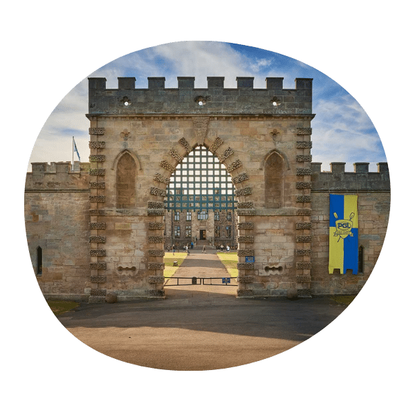 A stone castle gate with an arched entrance, viewed from outside. A yellow and blue flag hangs on the right wall; a courtyard and building are visible inside.