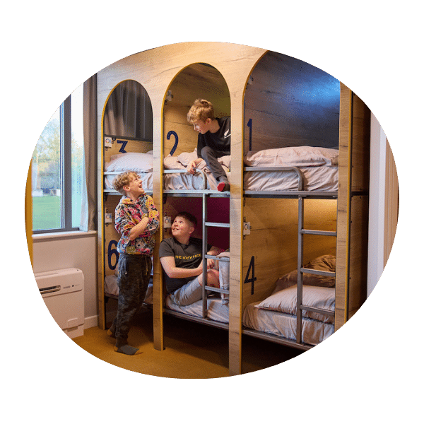 Three children interact while sitting and standing on two levels of numbered bunk beds in a brightly lit room with a large window.