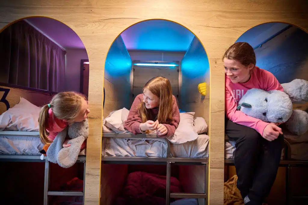 Three girls with stuffed animals sit and talk in adjacent wooden bunk bed cubbies, each lit with colorful lights.
