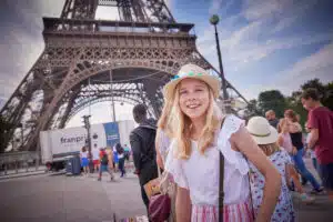 A girl in a hat smiles in front of the Eiffel Tower in Paris, with people walking and a truck in the background.