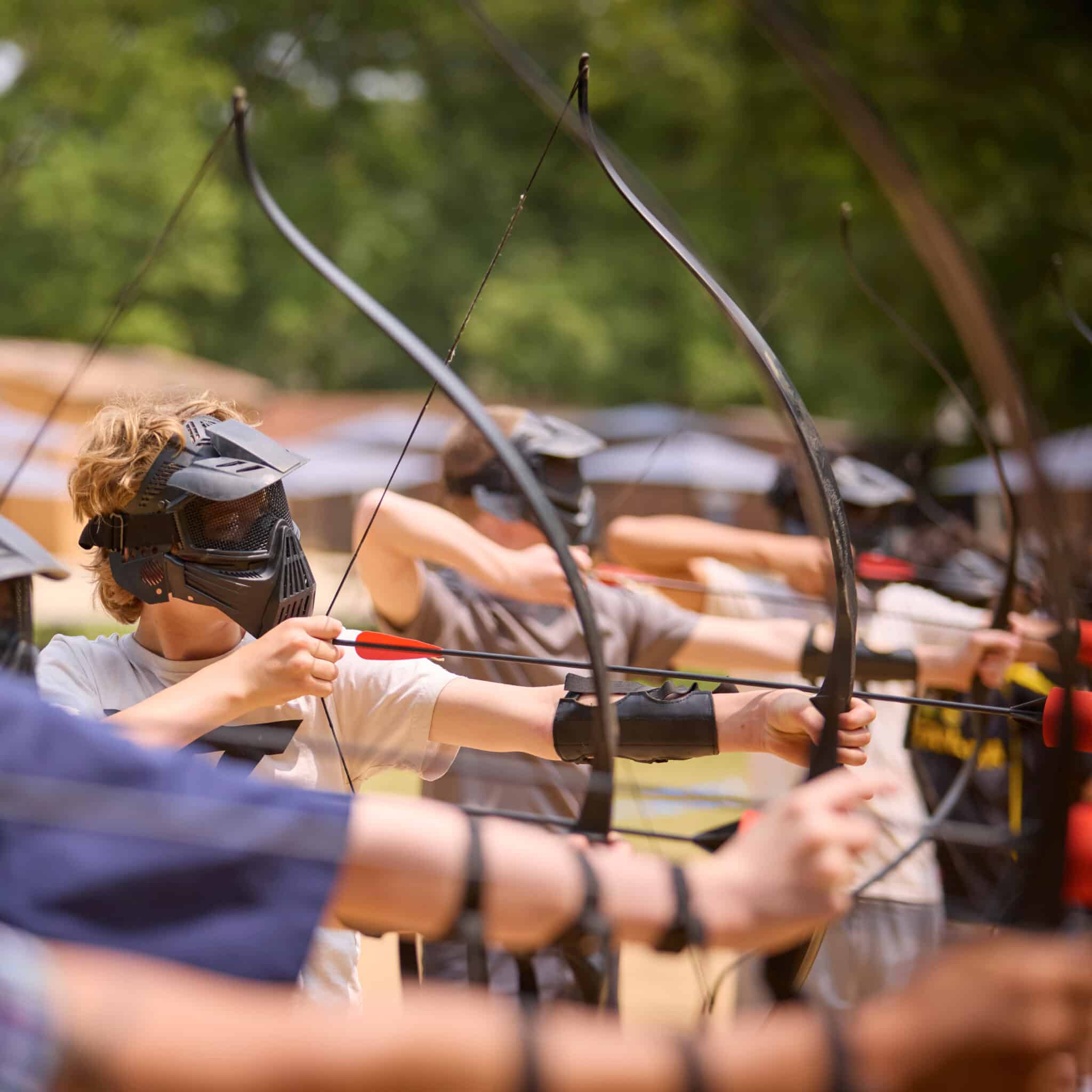 A group of people wearing protective masks and arm guards aim bows and arrows outdoors in a line, preparing to shoot.