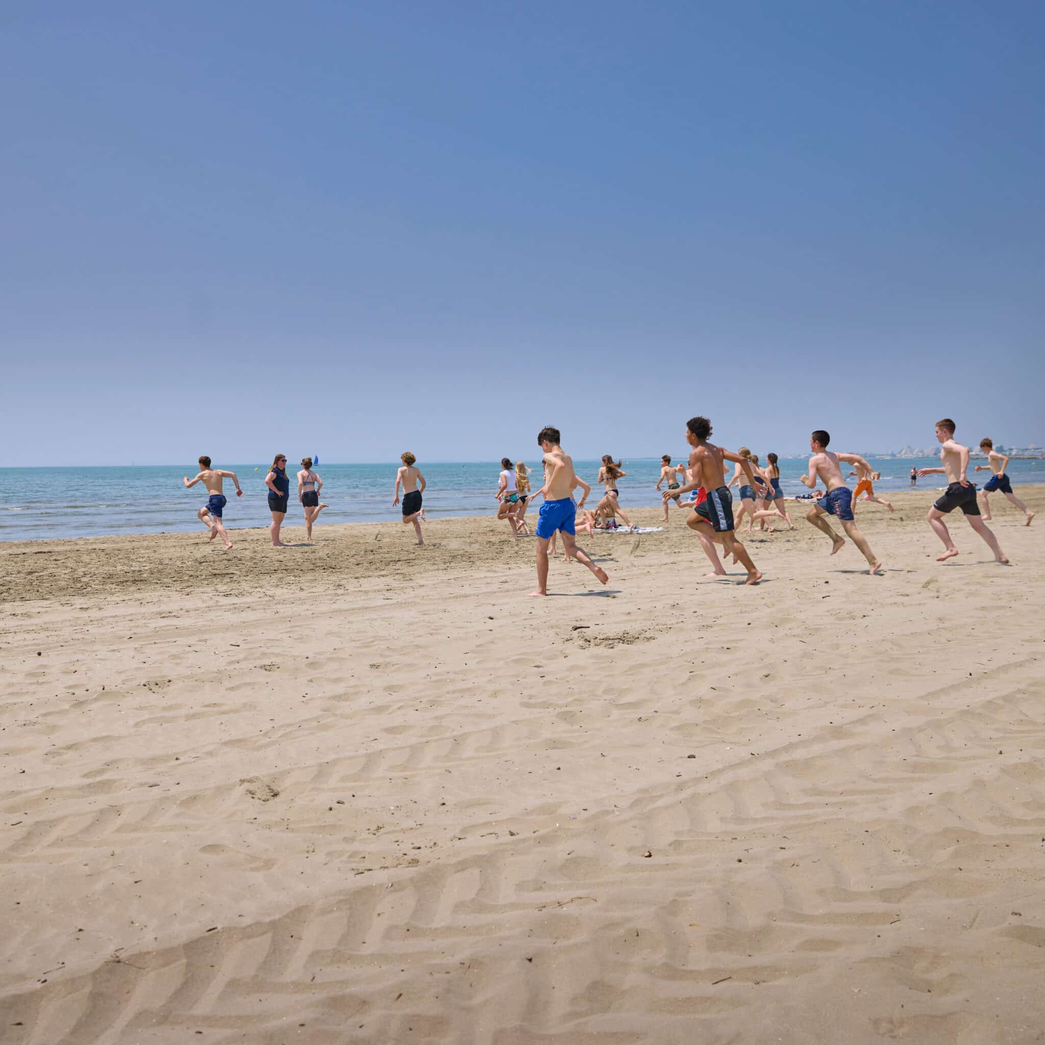 A group of people in swimsuits run toward the water on a sandy beach under a clear blue sky.