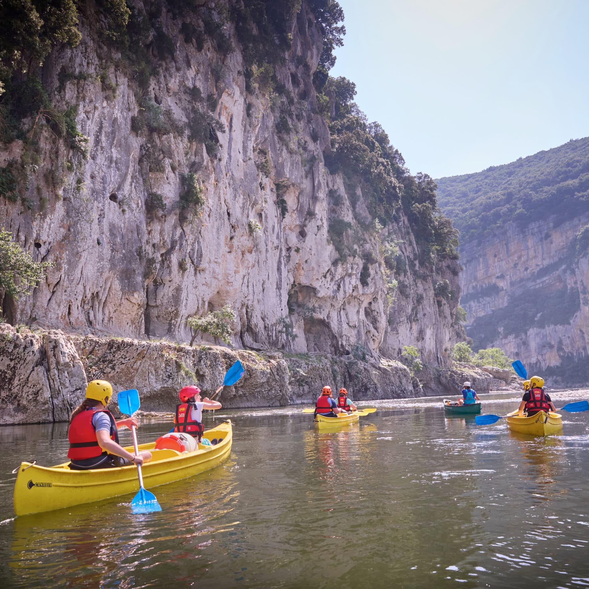 People in yellow kayaks paddle along a river bordered by steep rocky cliffs and greenery on a clear day.