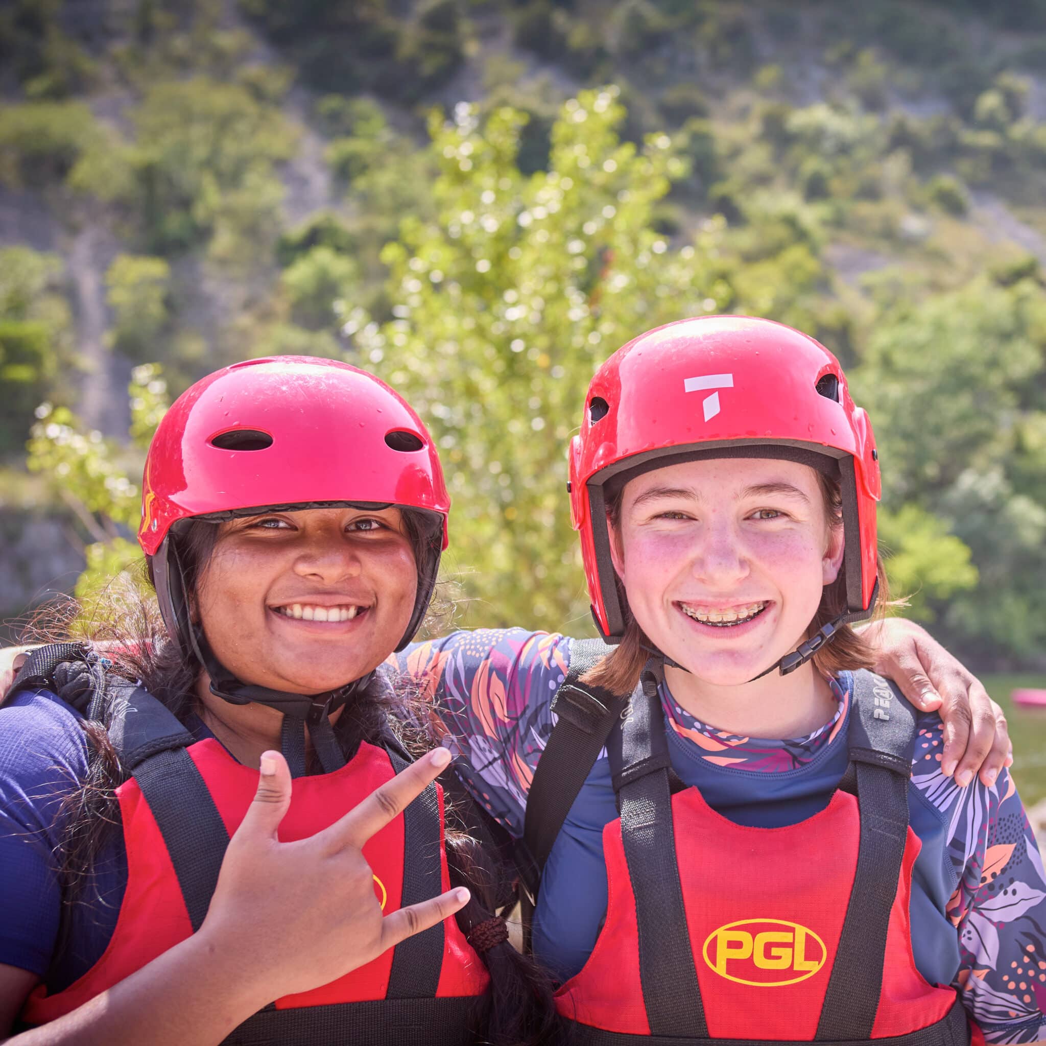Two people wearing red helmets and PGL life vests smile at the camera outdoors, with one person making a hand gesture. Trees and rocky terrain are visible in the background.