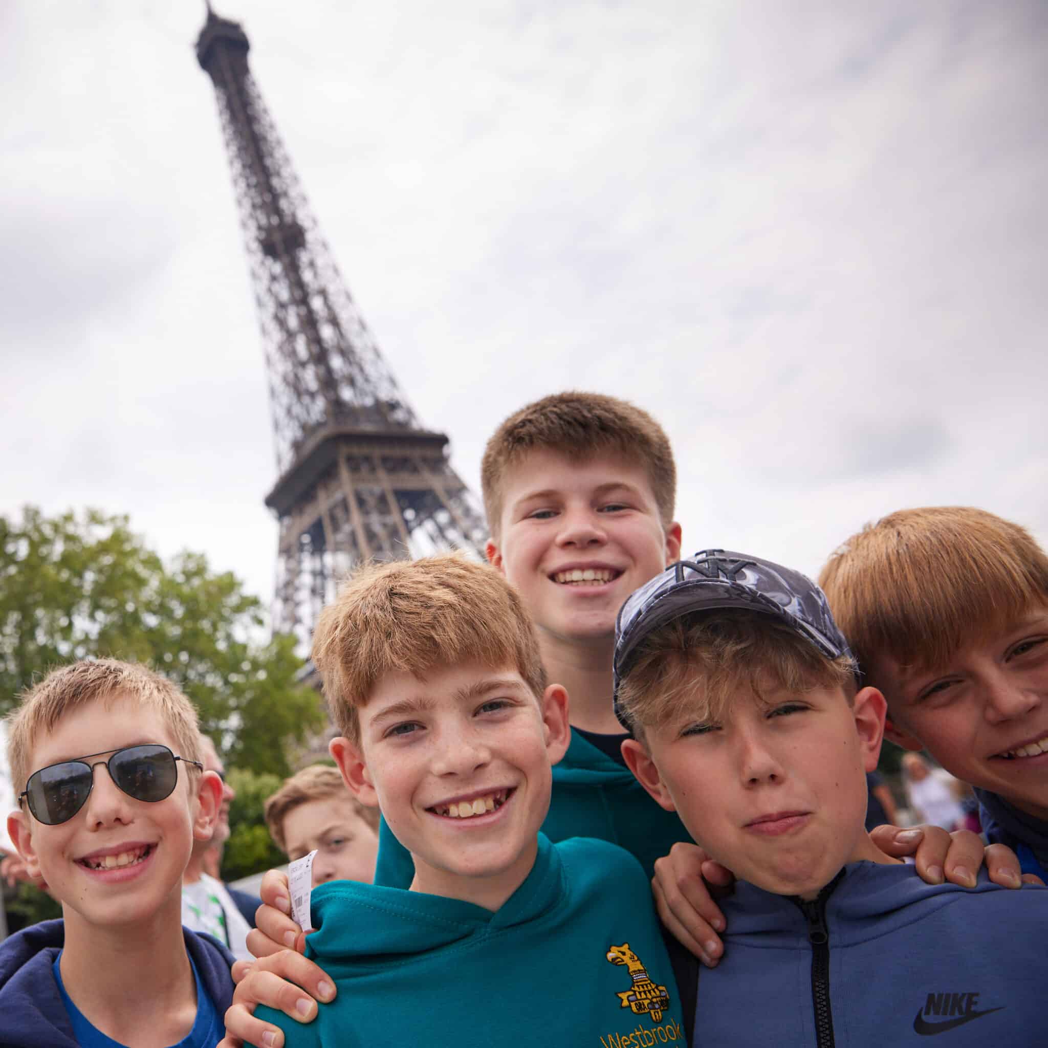 Five boys pose and smile in front of the Eiffel Tower on a cloudy day, with trees visible in the background.