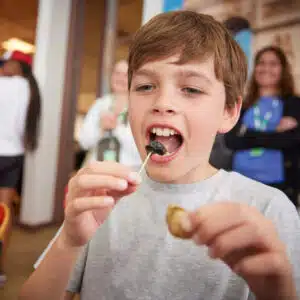 A boy in a gray shirt holds a snail shell in one hand and eats the snail with a toothpick.