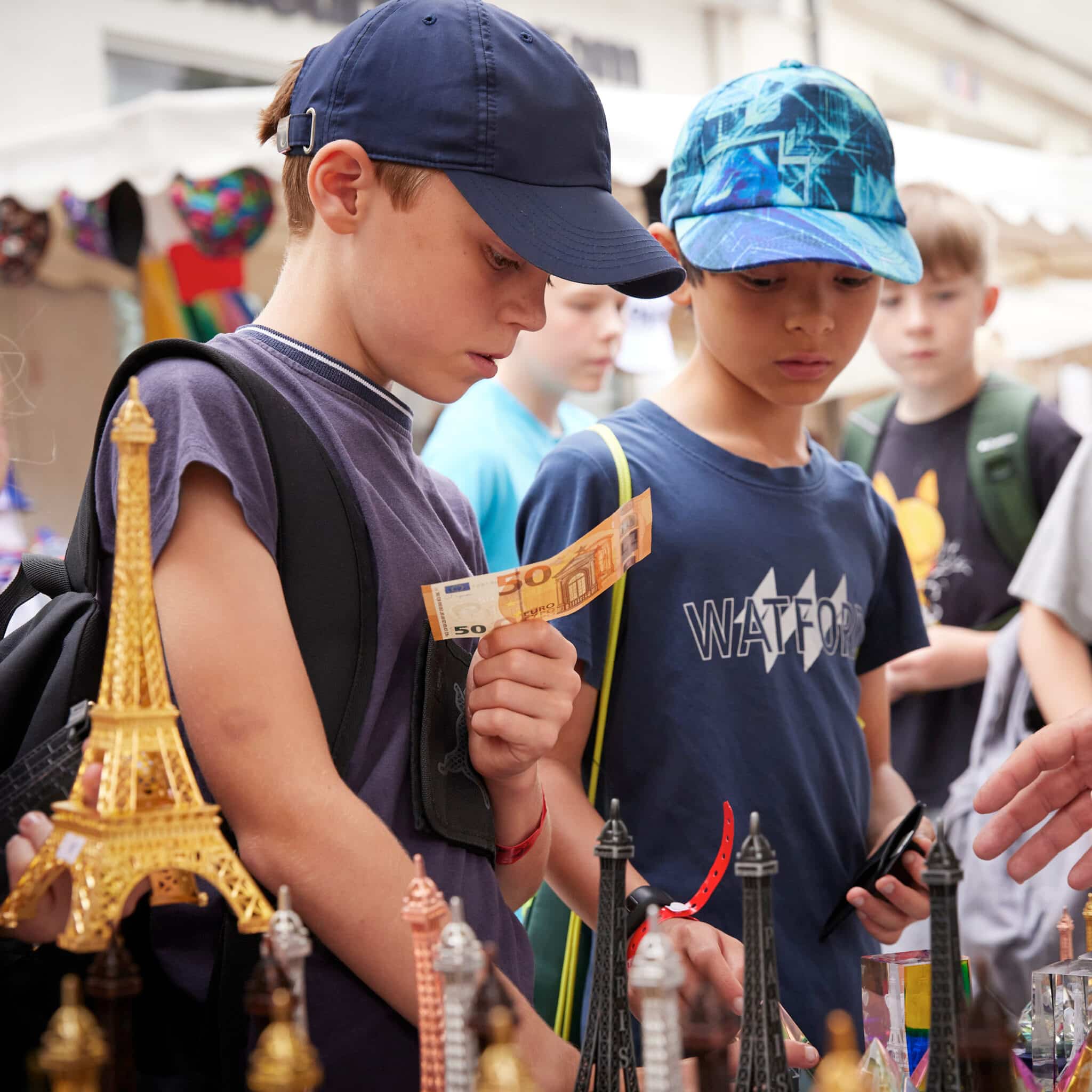 Two boys examine souvenirs at an outdoor market; one holds a 50 euro bill while the other looks at small Eiffel Tower models on the table.