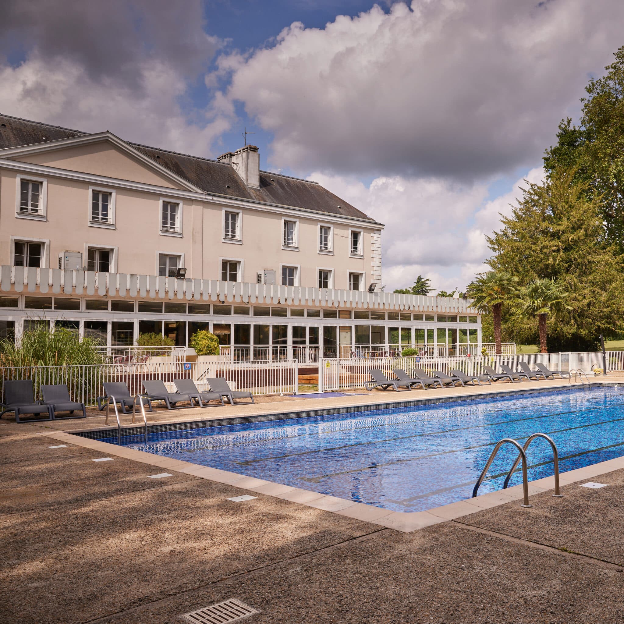Outdoor swimming pool with lounge chairs in front of a large beige building with multiple windows, under a partly cloudy sky.