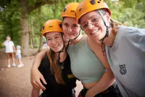 Three young people wearing orange helmets and casual clothing smile and pose together outdoors, with trees and blurred people in the background.
