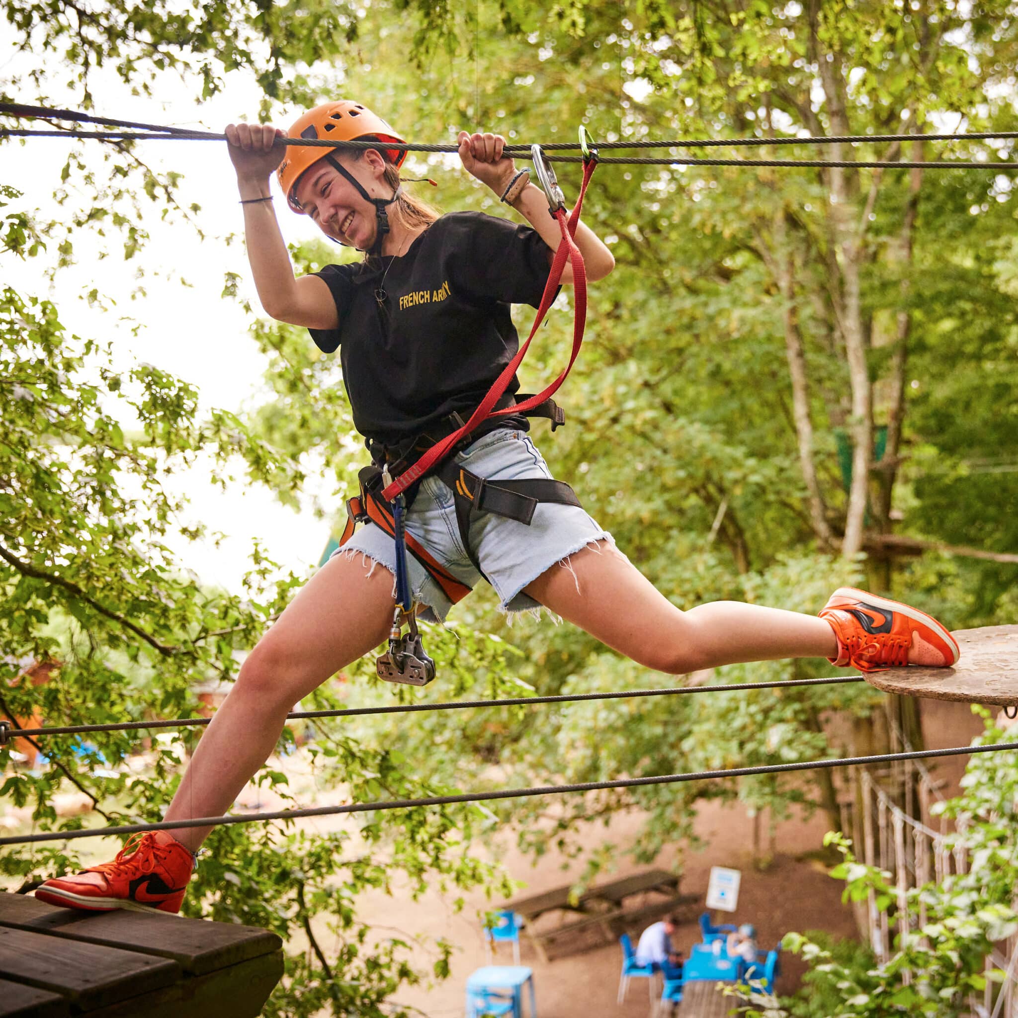 Person wearing a helmet and harness smiles while balancing between ropes on an outdoor adventure course in a forested area.