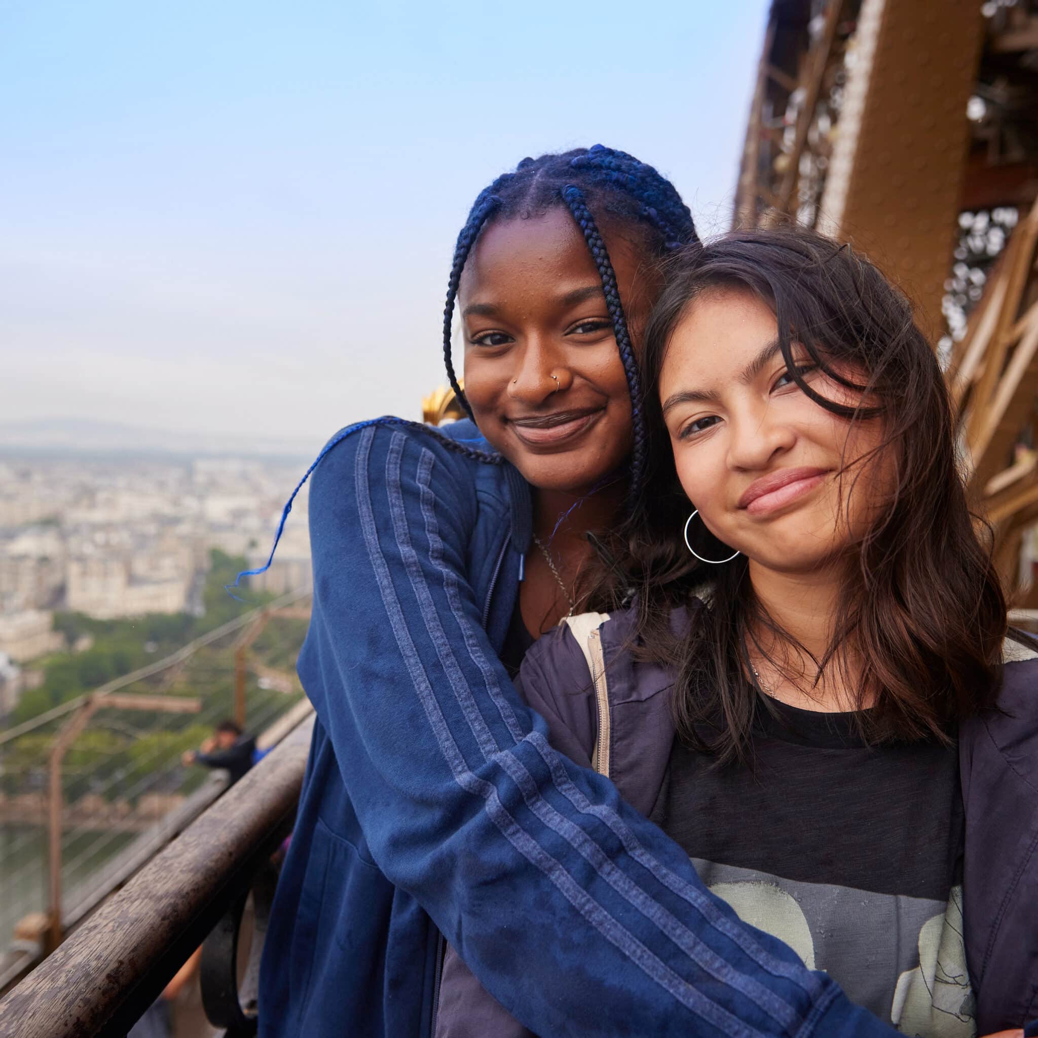 Two young women stand close together, smiling at the camera on an outdoor platform with a cityscape view in the background.