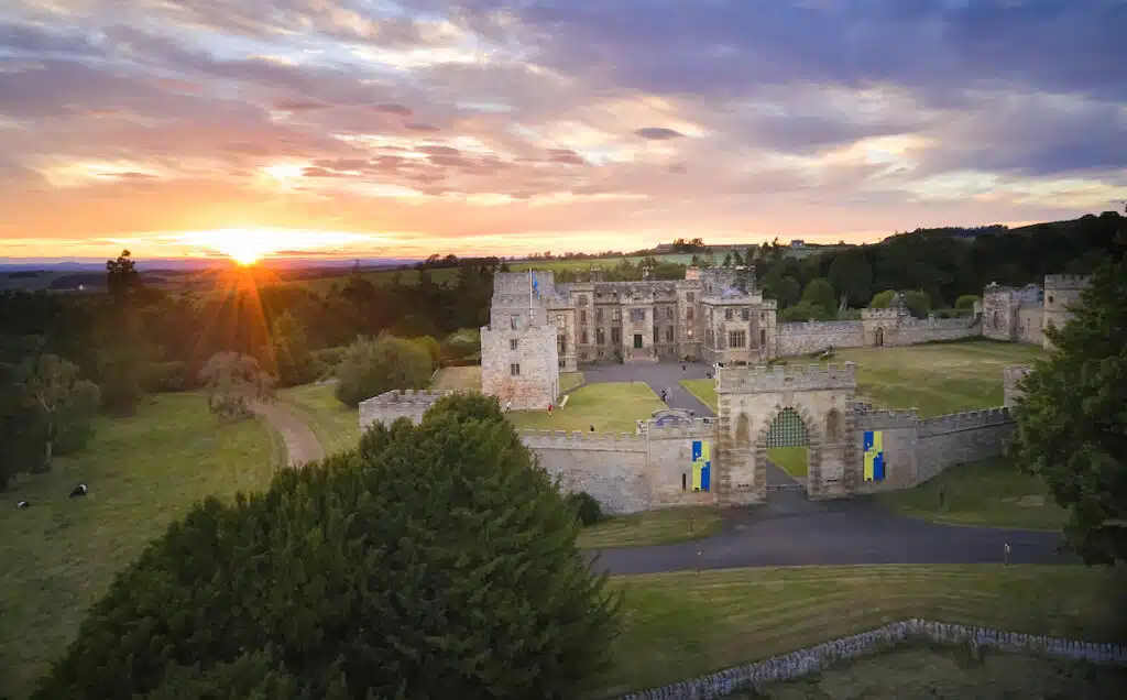A large stone castle with crenellated walls sits amid green fields at sunset; a gated entrance displays blue and yellow banners.