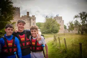Three young people wearing life jackets stand together outdoors, smiling, with a historic stone building and greenery in the background.