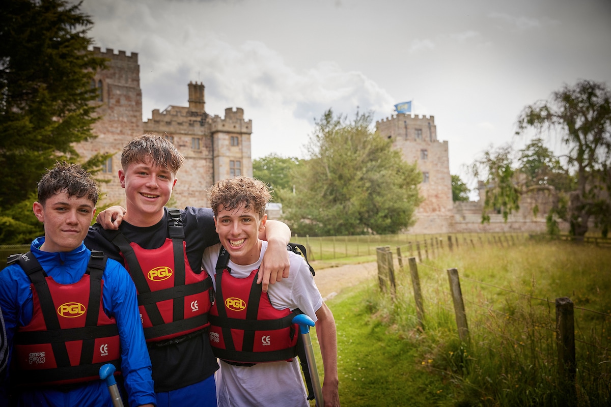 Three young people wearing life jackets stand together outdoors, smiling, with a historic stone building and greenery in the background.