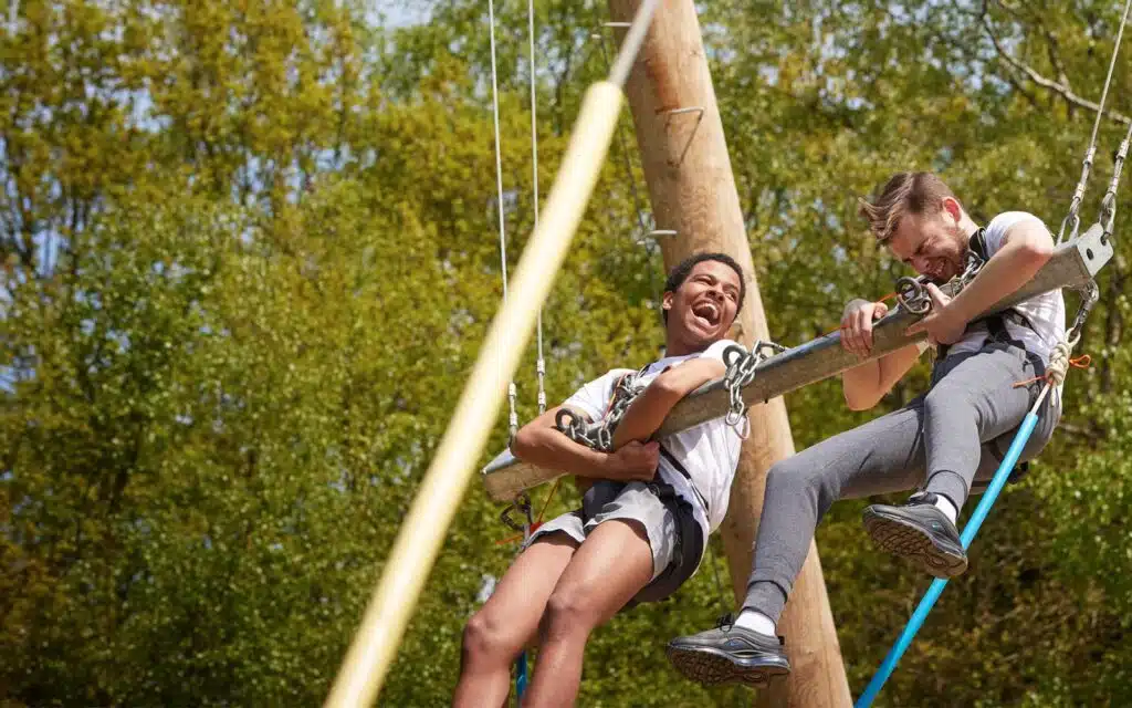 Two people wearing harnesses are suspended from a large swing outdoors, holding onto a metal bar, with trees and blue sky in the background.