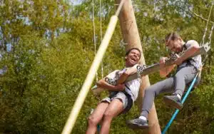 Two people wearing harnesses are suspended from a large swing outdoors, holding onto a metal bar, with trees and blue sky in the background.