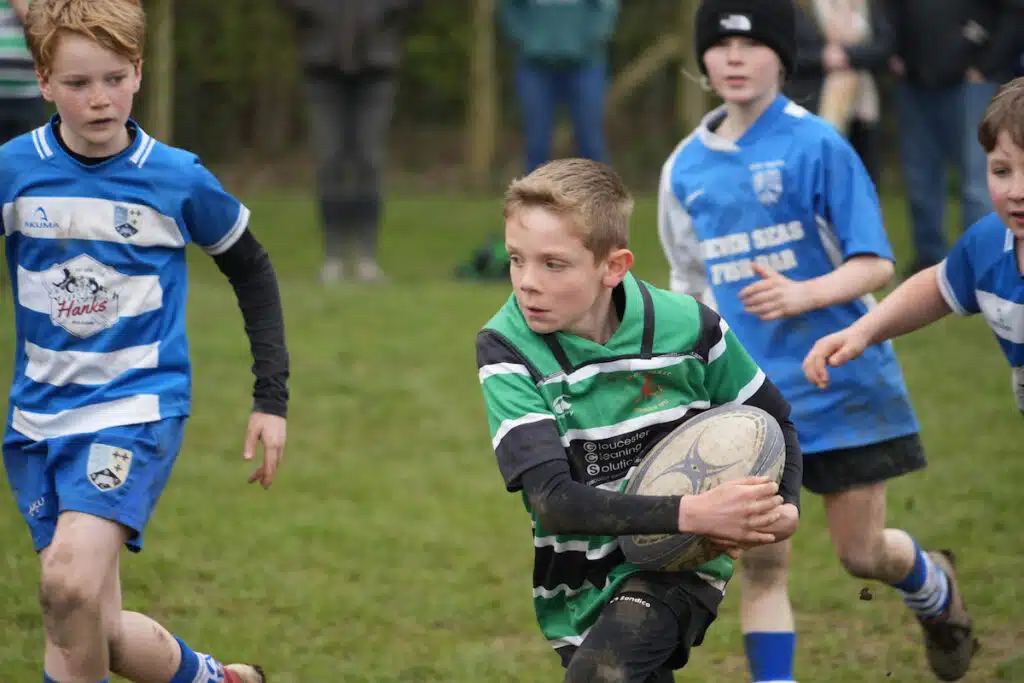 A boy in a green and black rugby jersey runs with the ball, pursued by boys in blue and white jerseys on a grassy field.