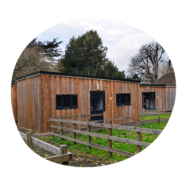 A single-story wooden building with a flat roof, black-framed windows, and a fenced grassy area in front, surrounded by trees.