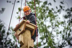 A child wearing a helmet and harness climbs a tall wooden structure outdoors, holding onto the top and smiling, with trees visible in the background.