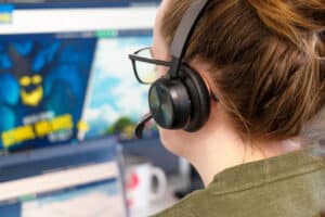 A person wearing a headset and glasses looks at a computer screen displaying a game or animated scene with a blue creature and text.