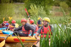 A group of people wearing helmets and life jackets sit in kayaks on a river, surrounded by green vegetation, preparing to paddle.