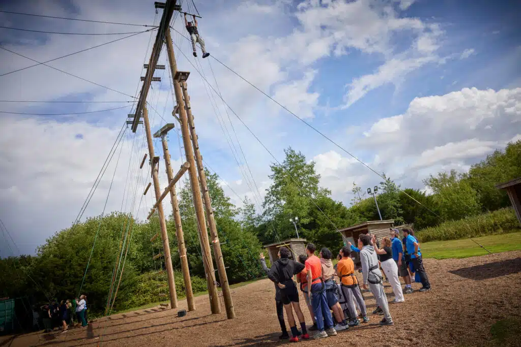 A group of people wearing harnesses watch as an individual climbs a tall wooden ropes course outdoors under a partly cloudy sky.