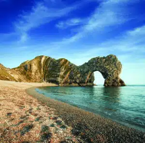 Natural rock arch known as Durdle Door stands on a pebbled beach along the coast, where blue sky and shallow waves invite a multi-activity day by the seaside.