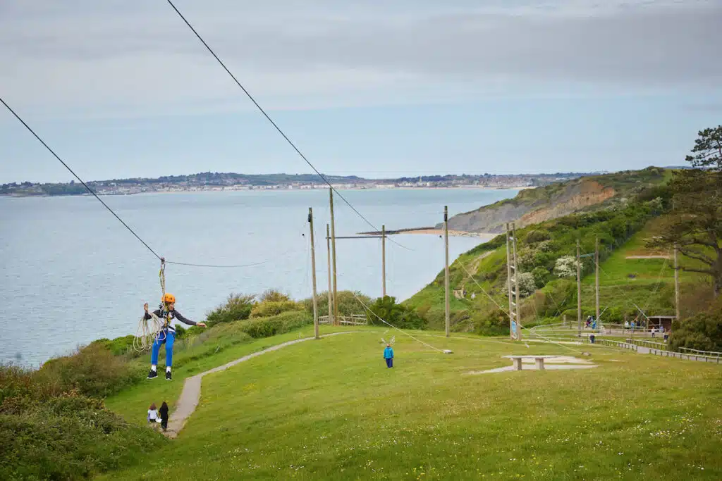 A person enjoys a multi-activity adventure, zip-wiring over a grassy hill near the coast, with sea, cliffs, and scattered people visible in the background under a partly cloudy sky.
