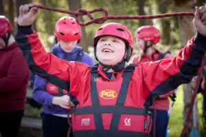 A child wearing a red helmet and outdoor gear holds up a climbing rope, ready for the send-trip, whilst others in similar attire stand in the background.