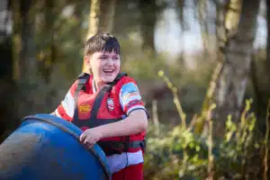A boy in a red sports shirt and life jacket smiles whilst holding a large blue plastic barrel outdoors, ready for a send-trip adventure, with trees in the background.