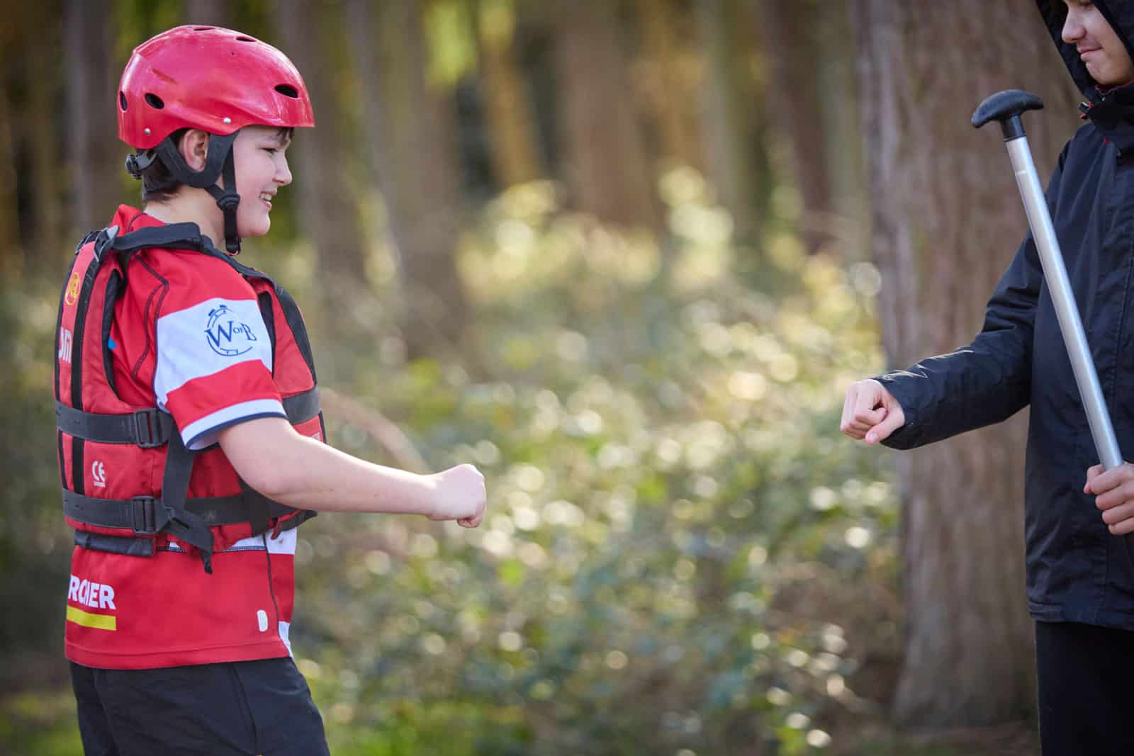 Two people playing rock-paper-scissors outdoors before their send-trip; one wears a red helmet and life vest, the other holds a paddle and sports a black jacket.