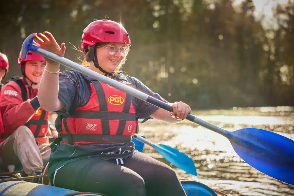 A person wearing a red helmet and life jacket is paddling a blue canoe on a sunlit lake, smiling and waving during their send-trip adventure.