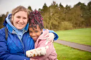 Two people stand outdoors on grass, smiling and hugging after a send-trip. One holds a water bottle. Trees and a paved path are visible in the background.