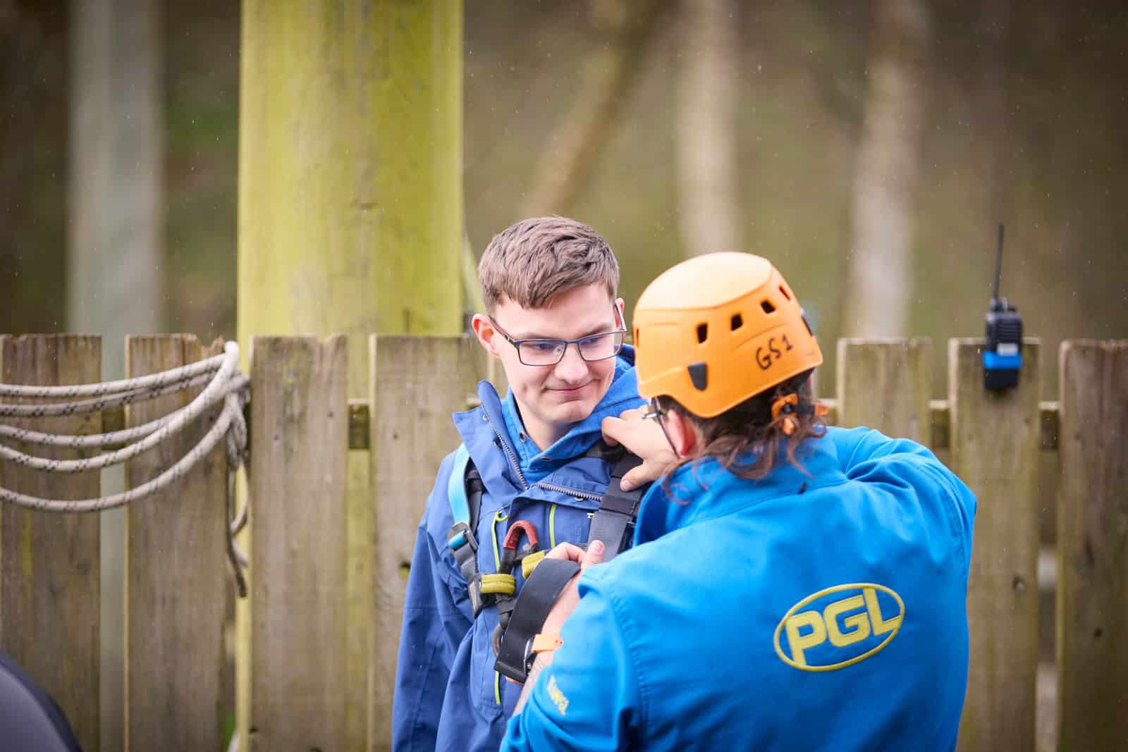 An instructor wearing a helmet and PGL jacket helps a person in glasses adjust their outdoor safety harness near a wooden fence, preparing them for their send-trip adventure.