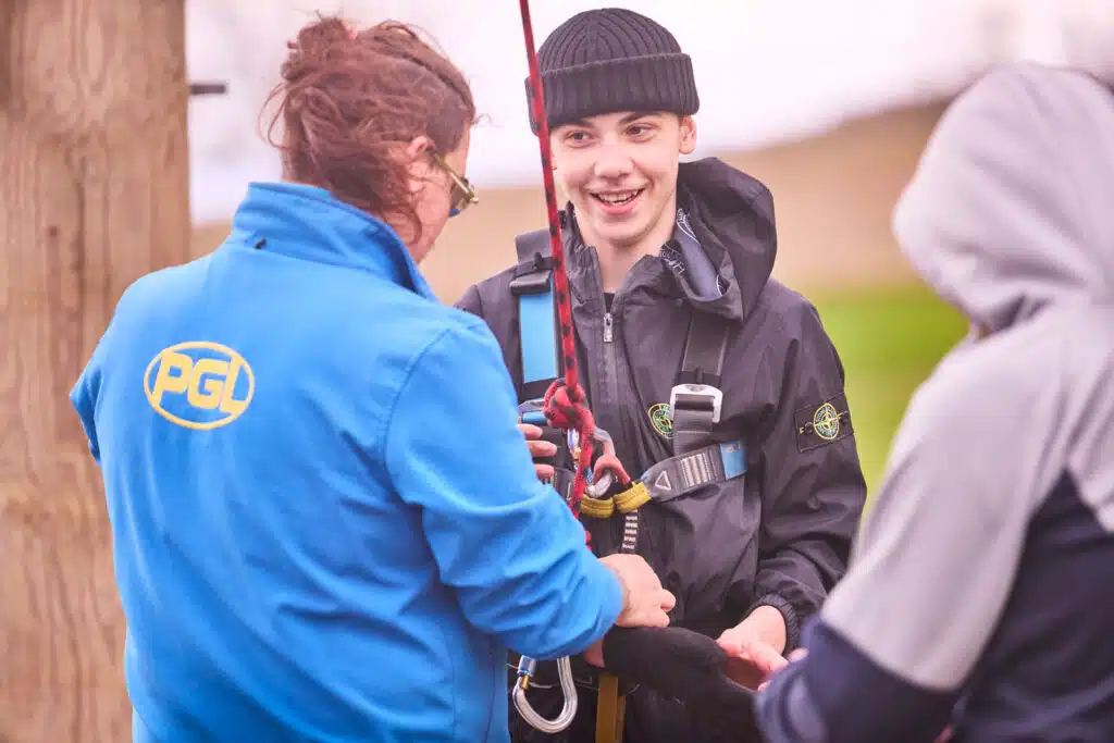 Three people are outdoors; one in a blue PGD jacket helps a smiling teen in harness gear get ready for an abseil, while another stands nearby.