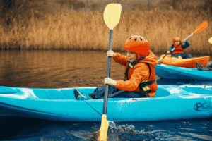 A child wearing a helmet and lifejacket paddles a blue kayak on a calm body of water, with other kayakers in the background.