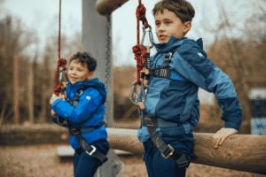 Two boys wearing blue jackets and harnesses stand next to wooden beams, holding onto ropes at an outdoor adventure park.