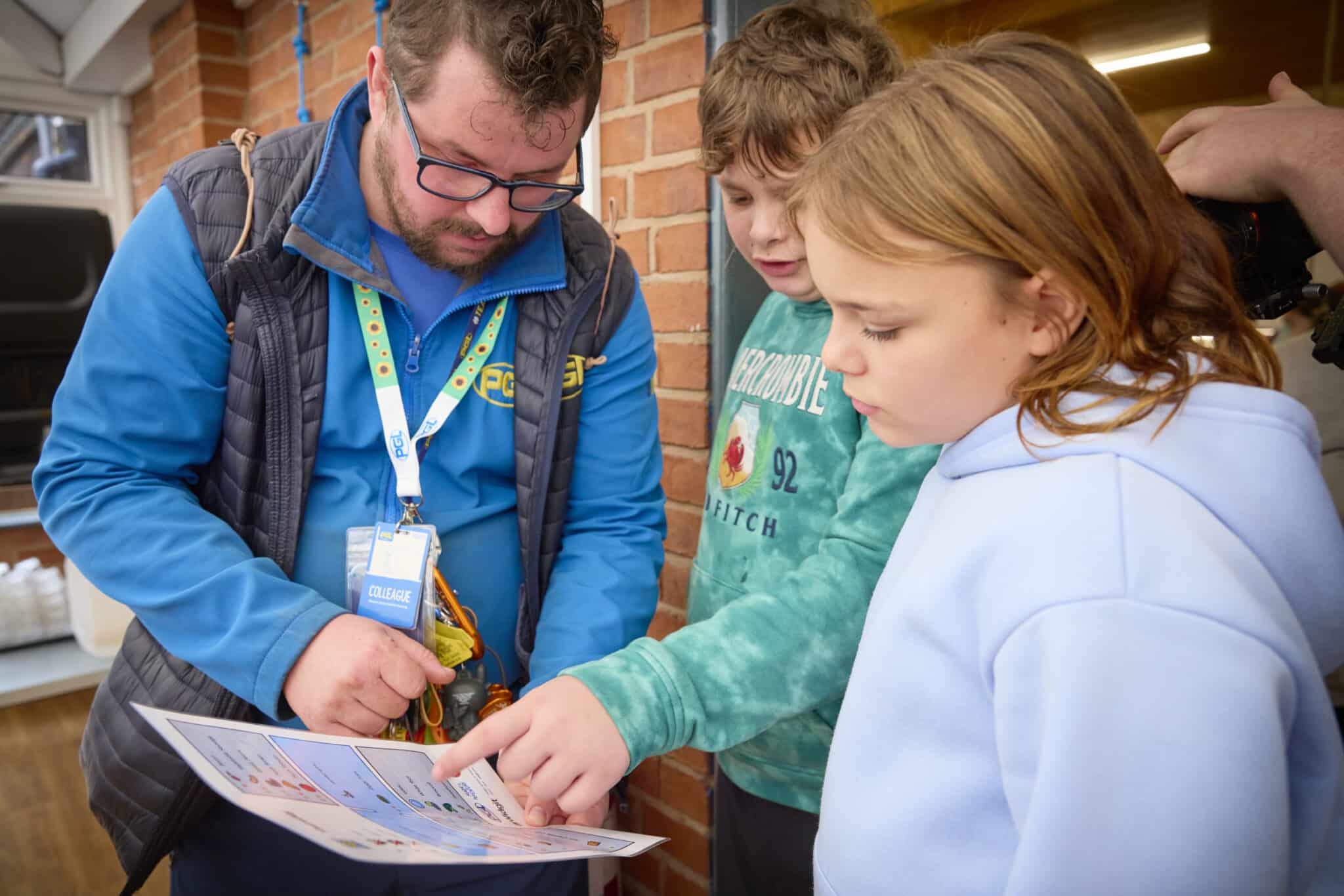 An adult shows two children a printed sheet with images and text, as the children look and point at the send-trip guide indoors near a brick wall.
